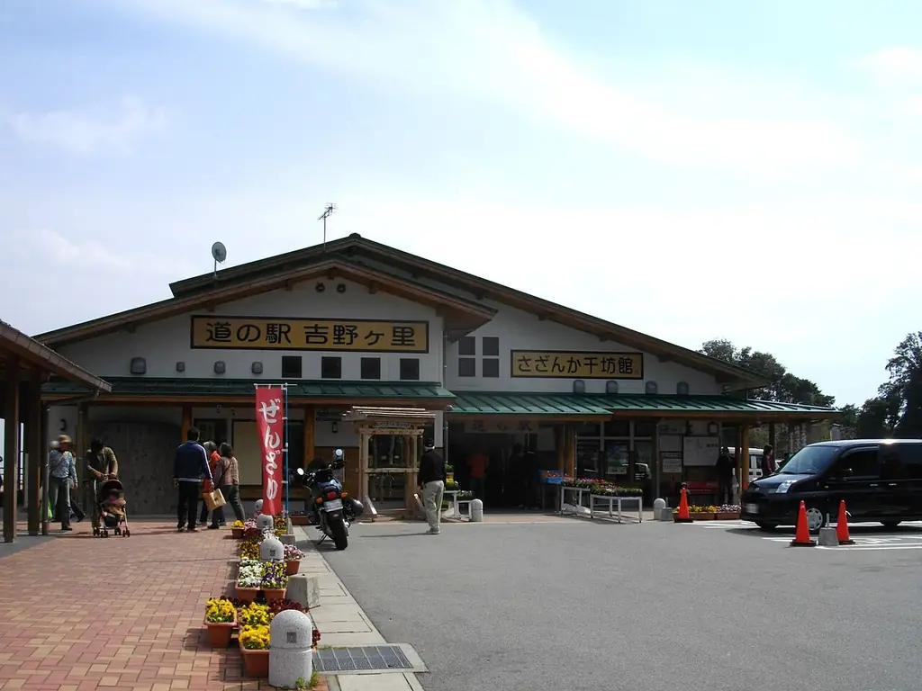 道の駅吉野ヶ里（吉野ヶ里町）の、「道の駅 吉野ヶ里」の看板が付いた建物と前を歩く人たちと花の鉢の写真