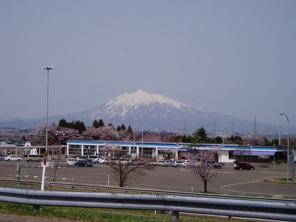 津軽サービスエリアの、広い駐車場と横に長い建物、桜の木と、うしろの雪の残る大きな山の写真