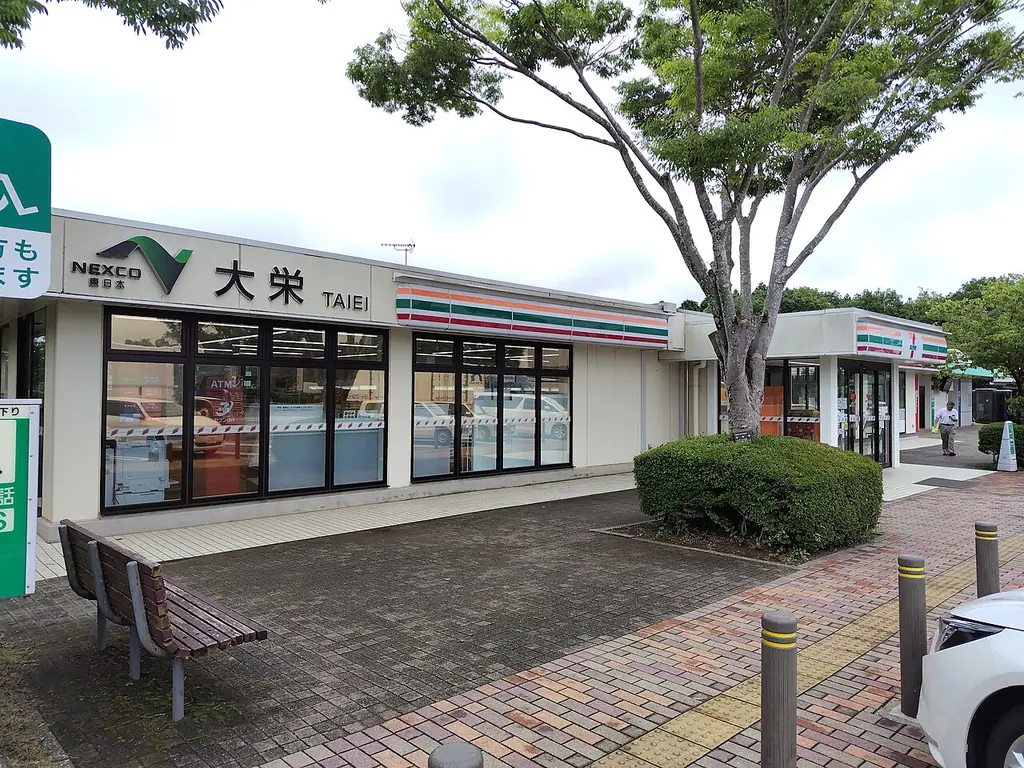 大栄パーキングエリアの、「大栄」の看板が付いた建物とコンビニの店先、前のベンチと木の写真