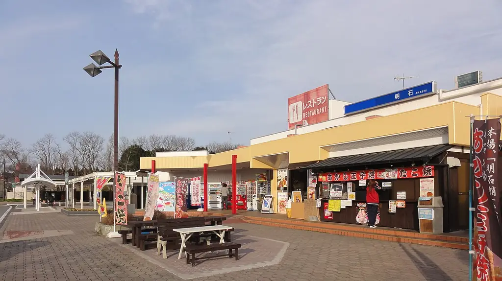 明石サービスエリアの、黄色い壁の店の建物とのぼり、前の広場に置かれたテーブルとベンチの写真