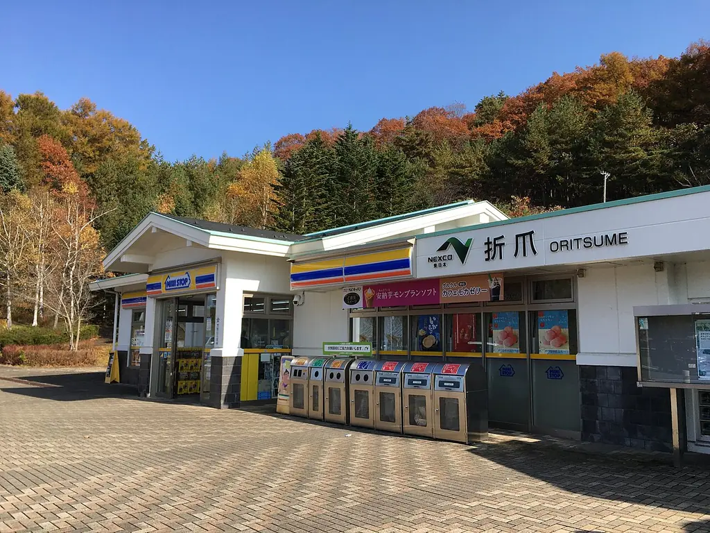 折爪サービスエリア（上り）の、「折爪」の看板が付いた建物とごみ箱の並ぶ店先、うしろの紅葉した山の写真