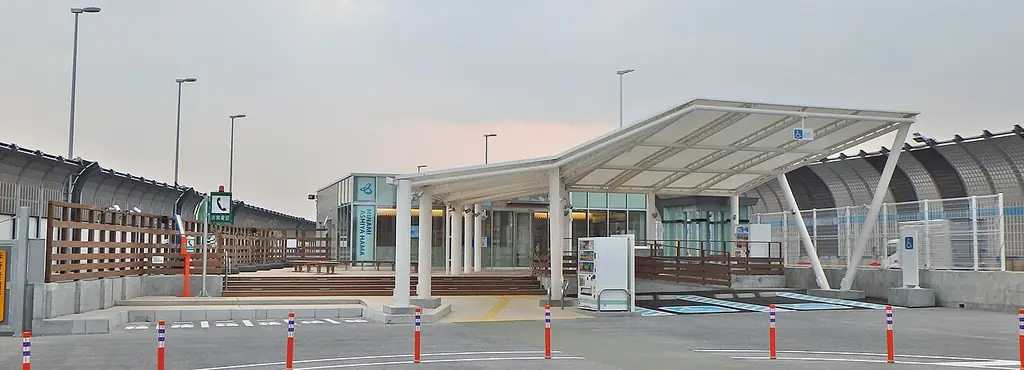 南芦屋浜パーキングエリアの、白い屋根の休憩所と木の柵、横に長い駐車場と照明柱の写真