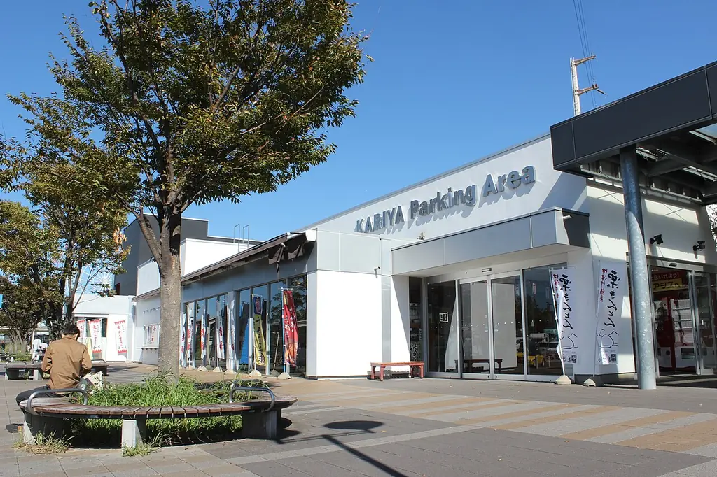 刈谷パーキングエリアの、「KARIYA Parking Area」の文字が付いた白い建物と、前の木とベンチの写真