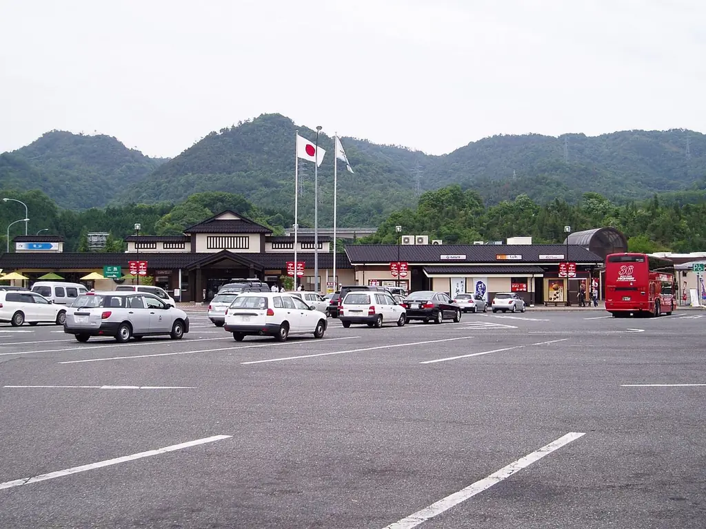 福山サービスエリア（山口方面）の、車が並ぶ広い駐車場と奥の建物、旗ざおと赤いバス、うしろの山並みの写真