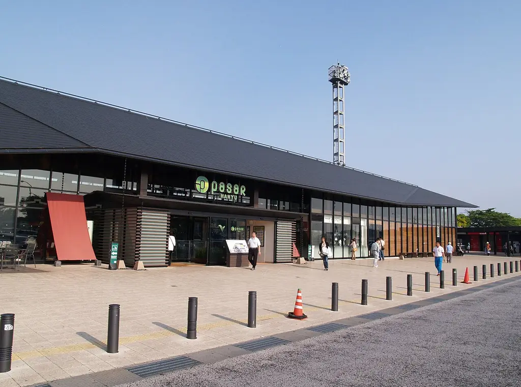 羽生パーキングエリア（下り）の、黒い大きな屋根の建物と照明の塔、前の歩道に並ぶ車止めと歩く人たちの写真