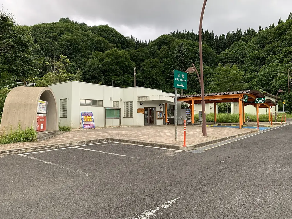 月山湖パーキングエリアの、半円の形の壁と白い建物、木の屋根の休憩所、うしろの緑の山の写真