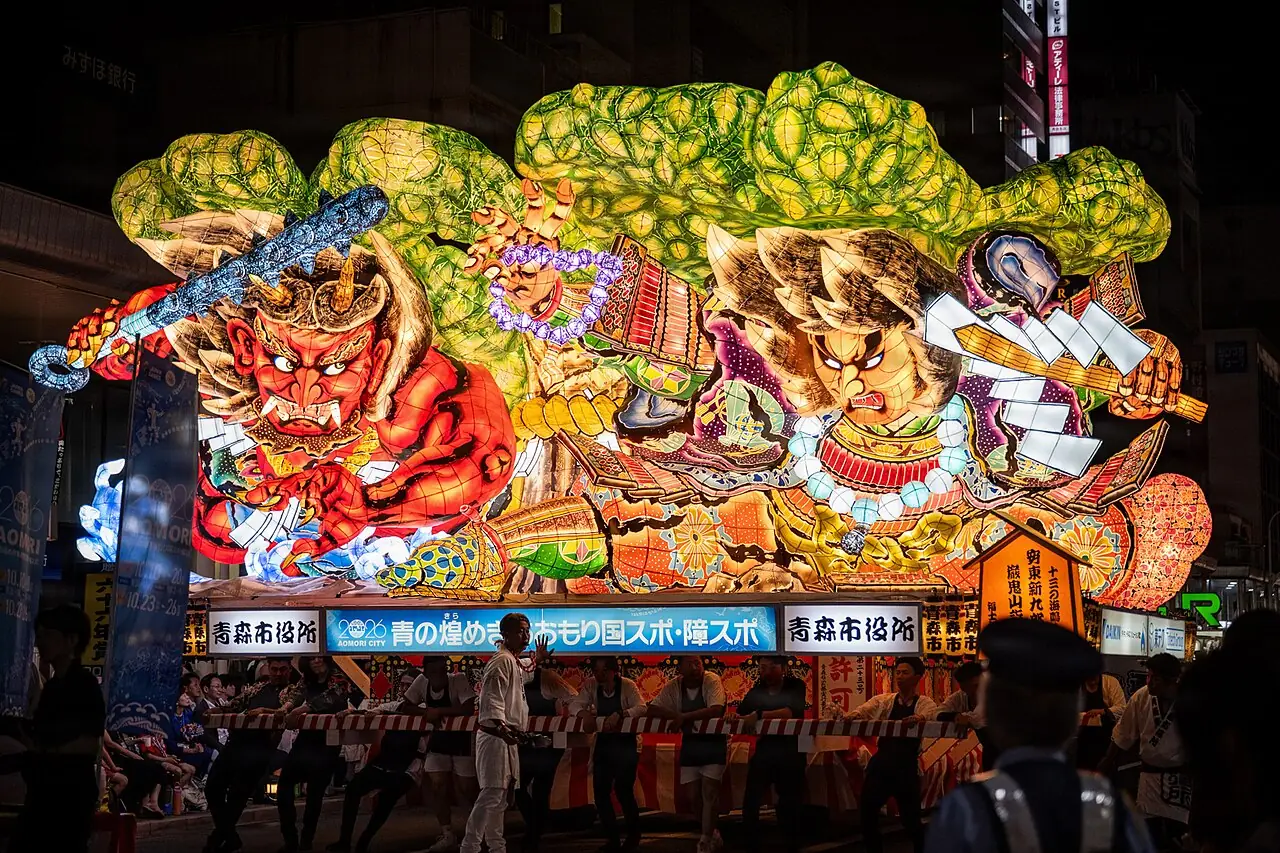 青森ねぶた祭で、武者などの絵が光る大きなねぶたが夜の通りを進み、まわりを人たちが囲む様子の写真