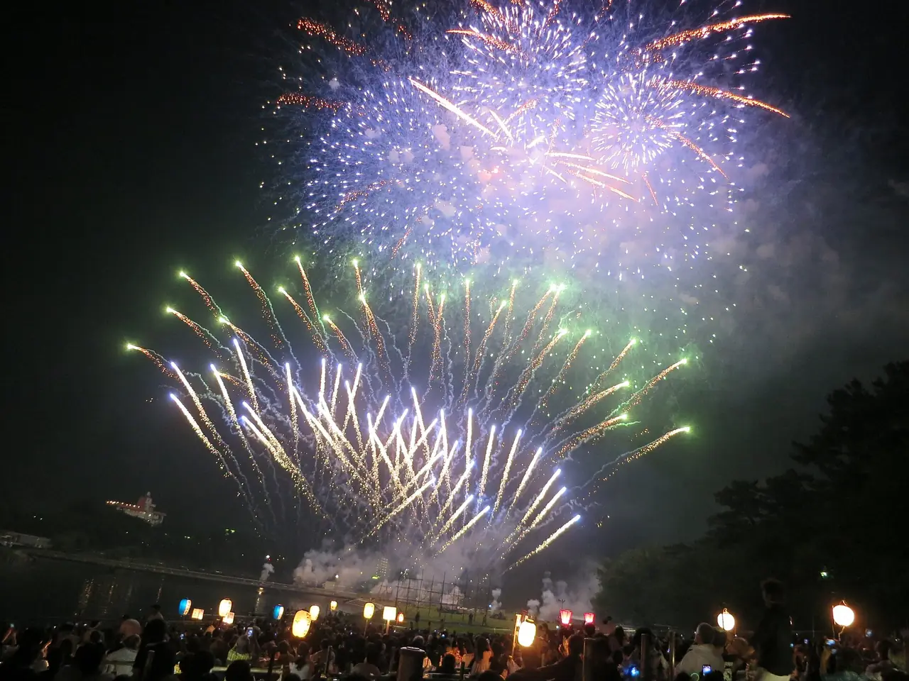 岡崎城下家康公夏まつり花火大会で、青白い花火と扇形に広がる金色や緑の花火が開き、下に提灯と見物の人たちが並ぶ様子の写真