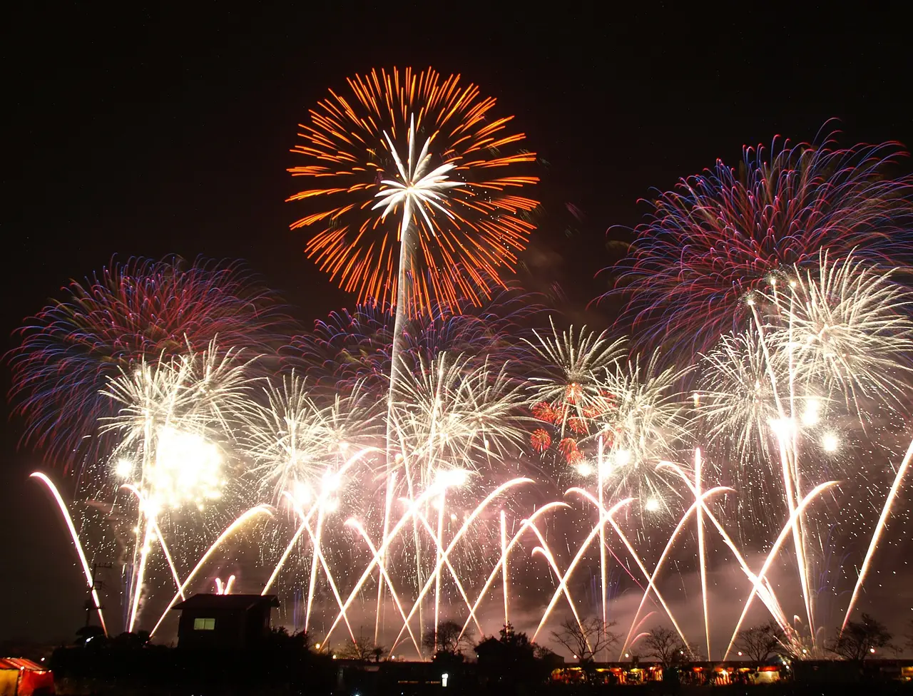 土浦全国花火競技大会で、夜空にオレンジや紫、白の花火がいくつも重なって開き、下に建物や木の影が並ぶ様子の写真