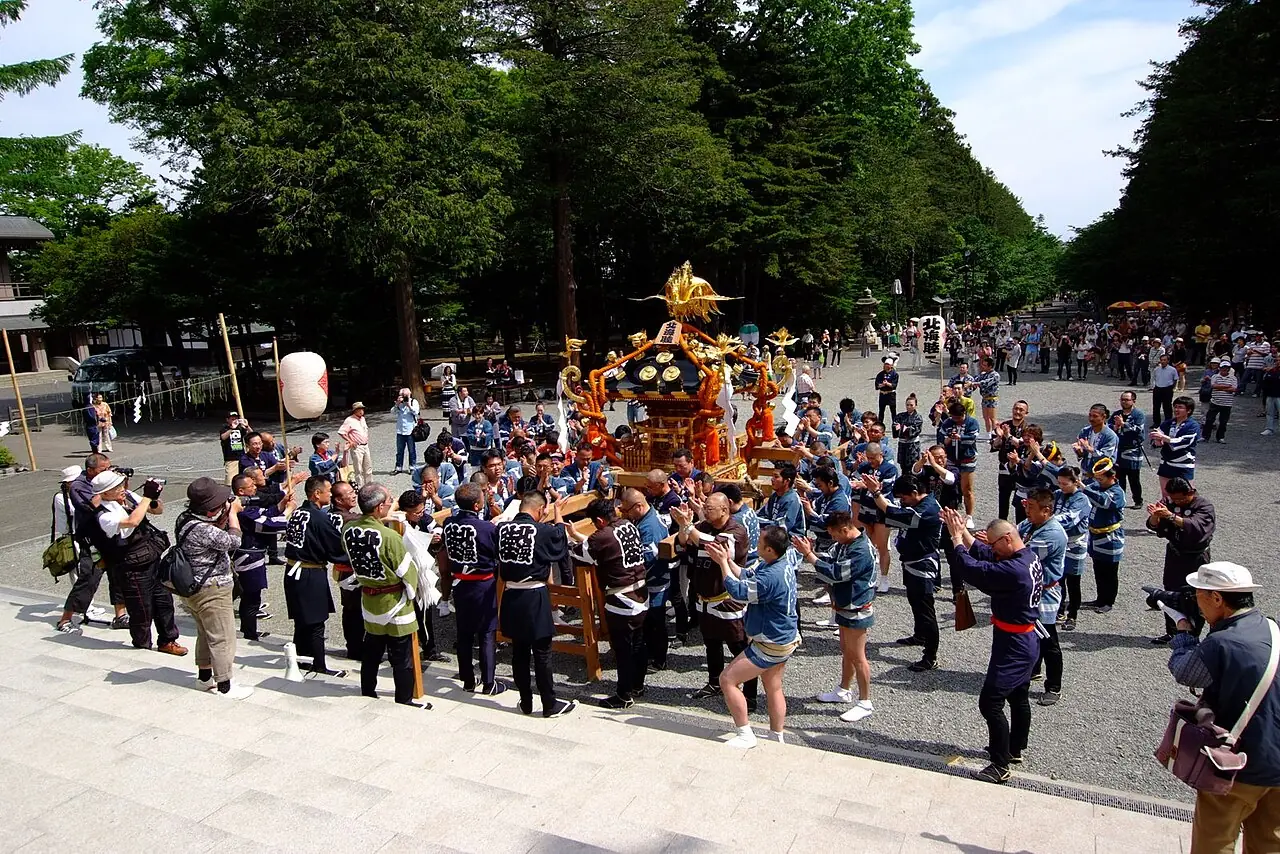 札幌まつり(北海道神宮例祭)で、木立の中の広い道に金色の神輿が出て、そろいの法被の人たちが囲む様子の写真