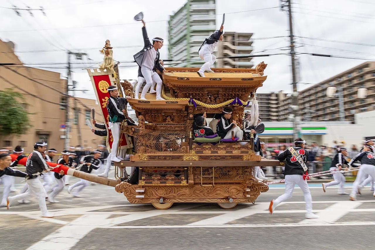 堺の津久野だんじり祭で、屋根に人が立つ彫り物のだんじりを、人たちが曳いて勢いよく走る様子の写真