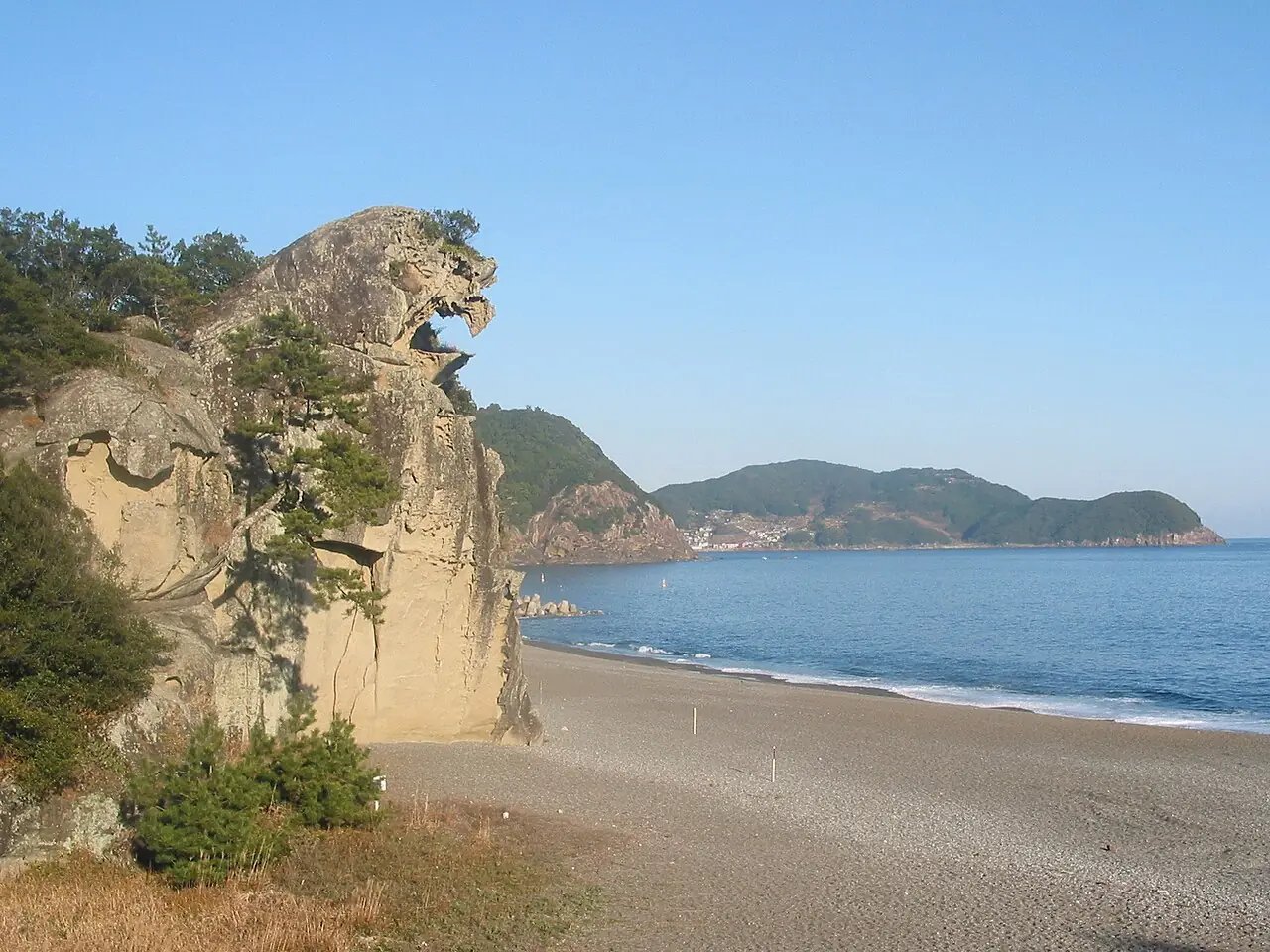 七里御浜海岸(会場)の、獅子の顔のような形の大きな岩と、海沿いに弓なりに続く浜と奥の町の写真