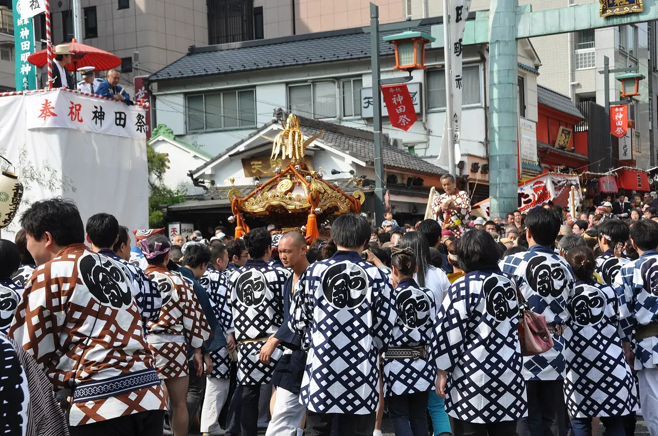神田祭で、そろいの法被の人たちが金色の神輿を囲み、横に祭りの幕、奥に門と提灯が見える様子の写真