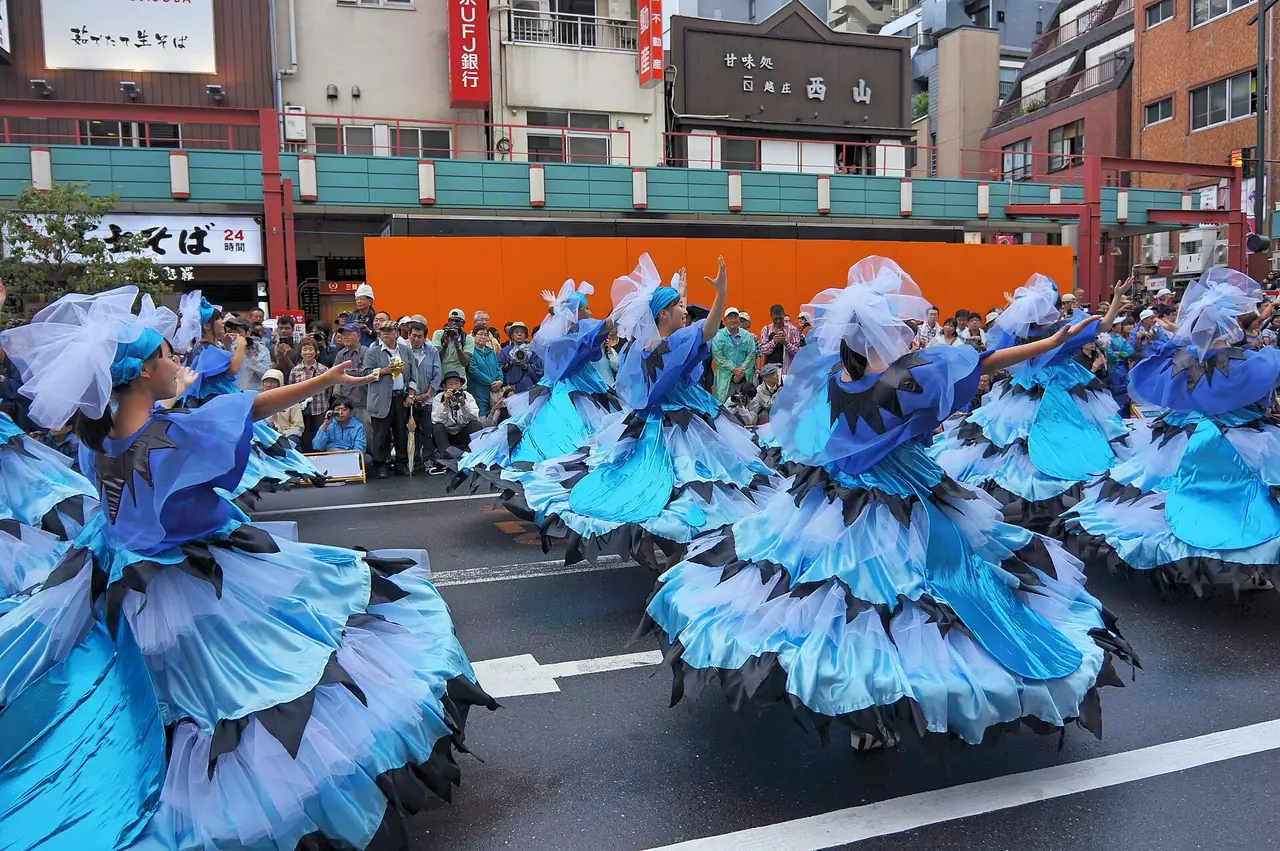浅草サンバカーニバルで、青と水色の大きく広がる衣装の踊り手たちが通りを進み、沿道に人たちが並ぶ様子の写真