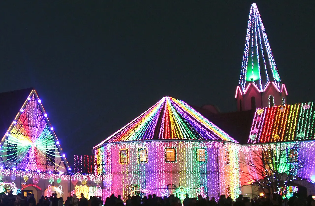 東京ドイツ村のイルミネーションで、三角屋根の建物やとがった塔が虹色の電飾に彩られ、手前に人たちの影が並ぶ様子の写真