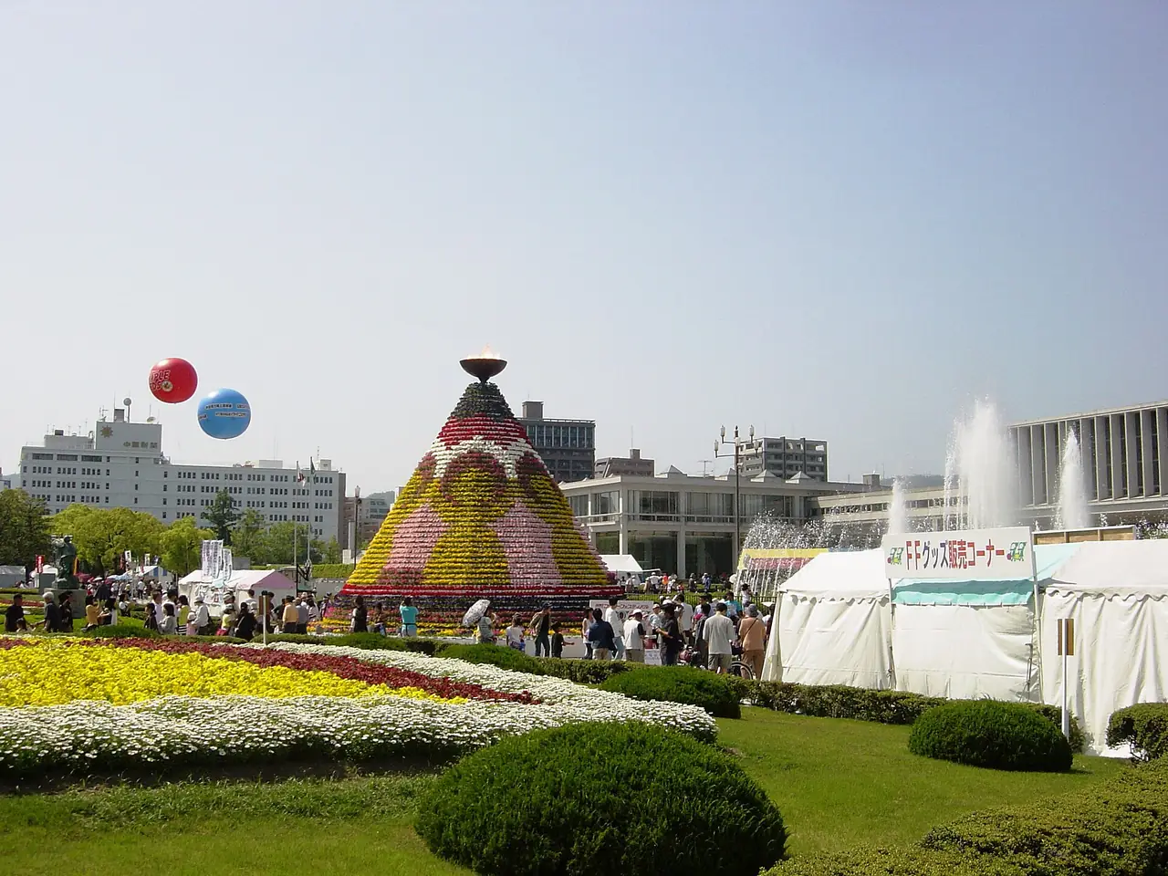 ひろしまフラワーフェスティバルで、花で飾った円すい形の塔と色とりどりの花壇、催しのテントが並ぶ広場の写真