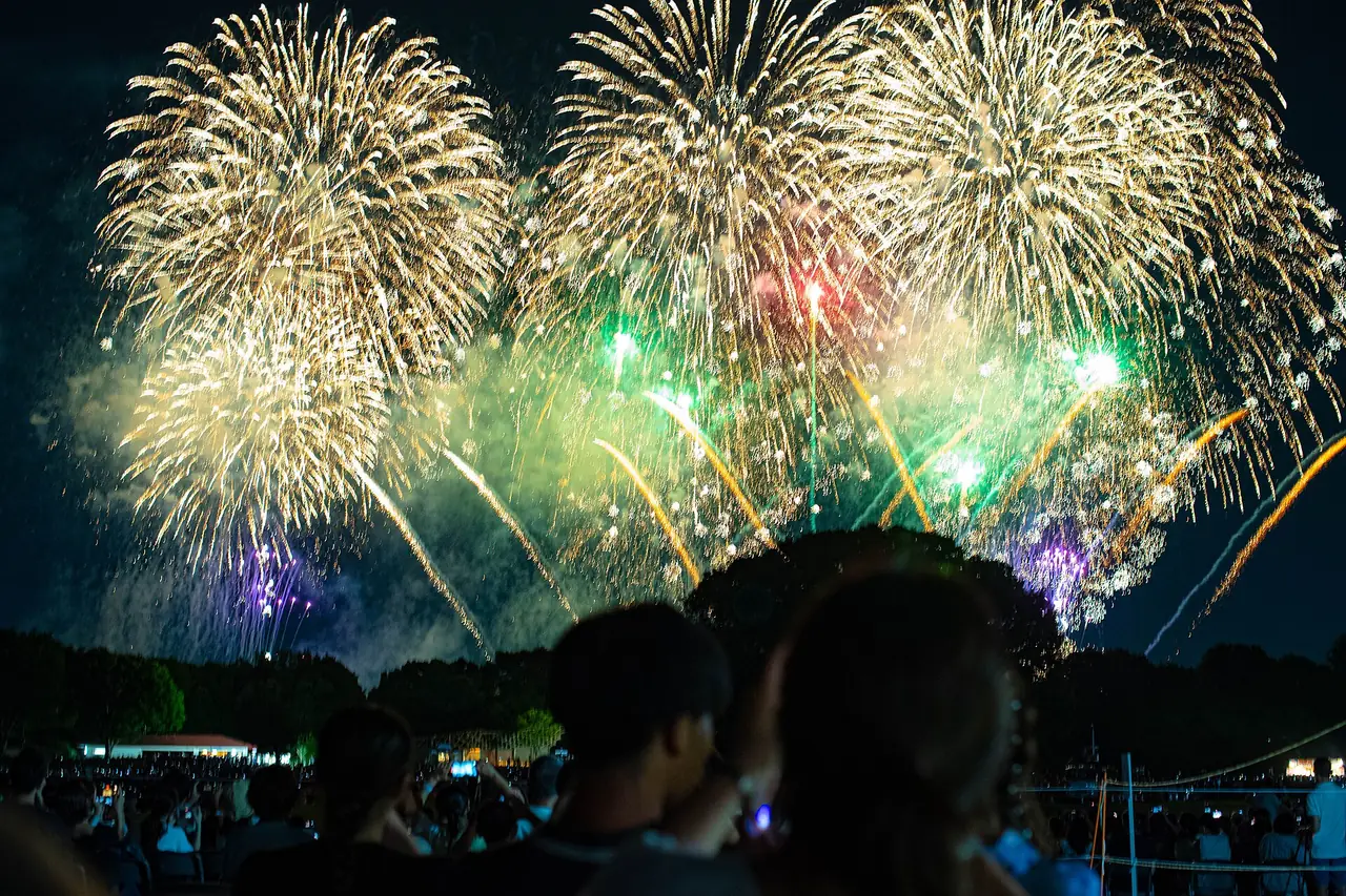 立川まつり国営昭和記念公園花火大会で、木立の上に金色や緑の花火がいくつも開き、見上げる人たちの影が見える様子の写真