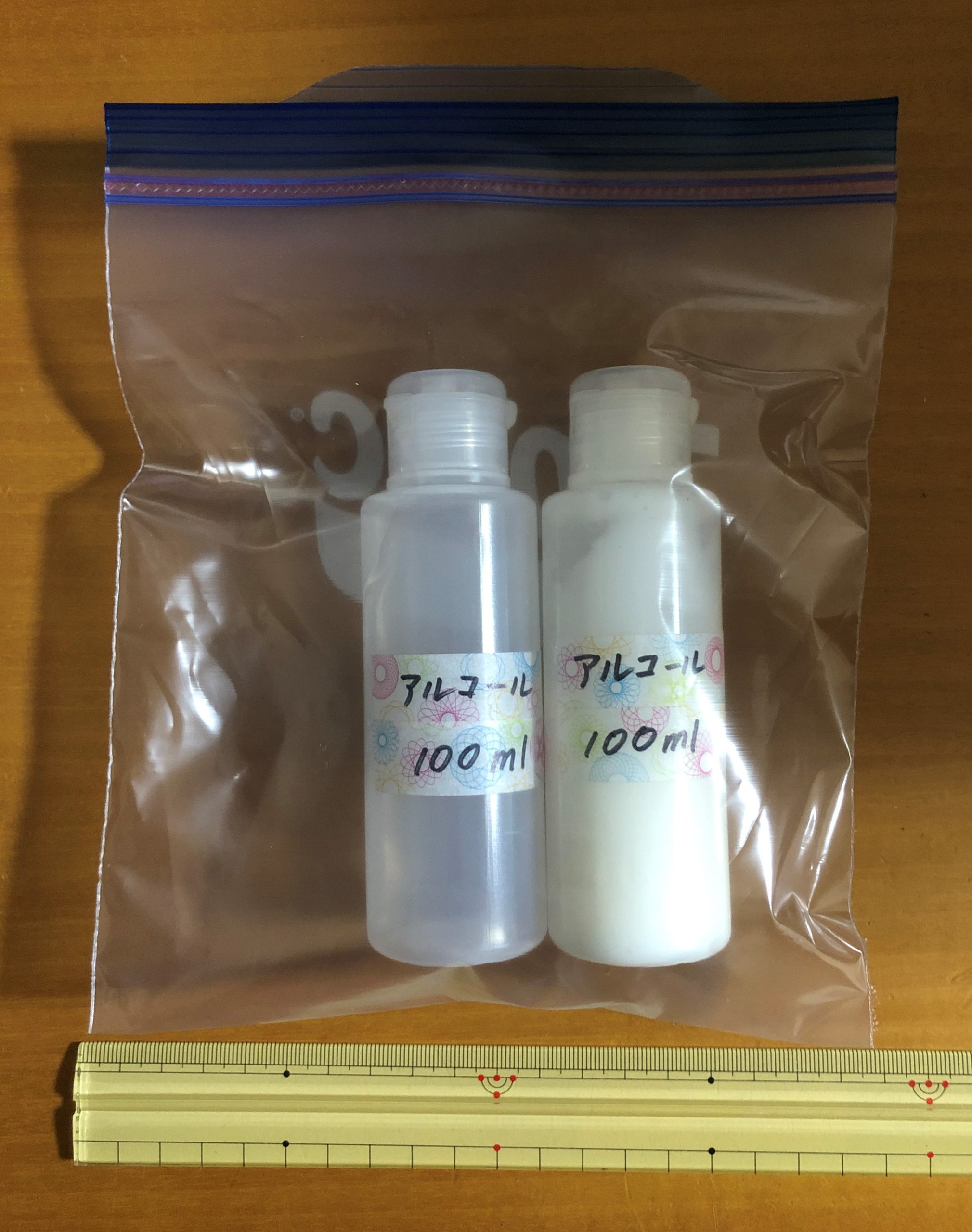 アルコール100mlと書いた容器2本をチャック付きの透明な袋に入れて定規と並べた写真