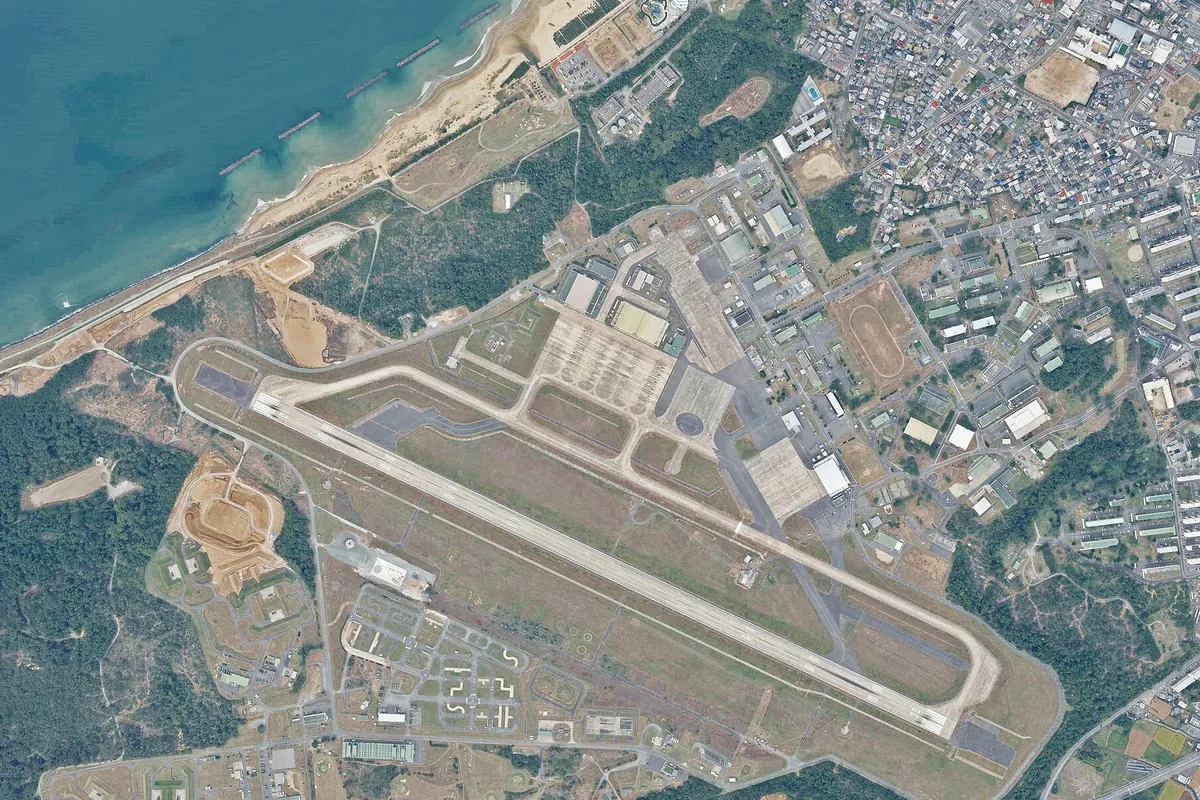 海沿いの砂浜近くに延びる滑走路と駐機場・建物が並ぶ航空基地を上空から写した写真