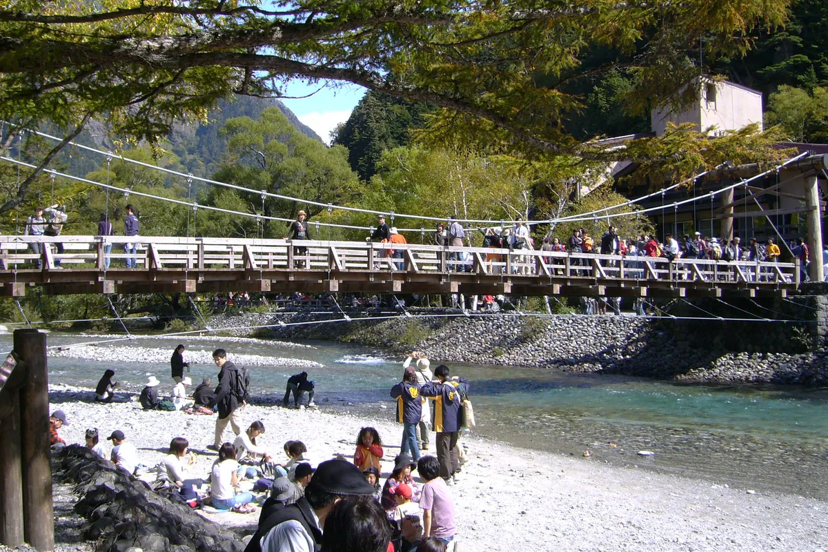 上高地の河童橋を観光客が渡り、川辺にも大勢の人がいる中で山並みと橋が写る写真