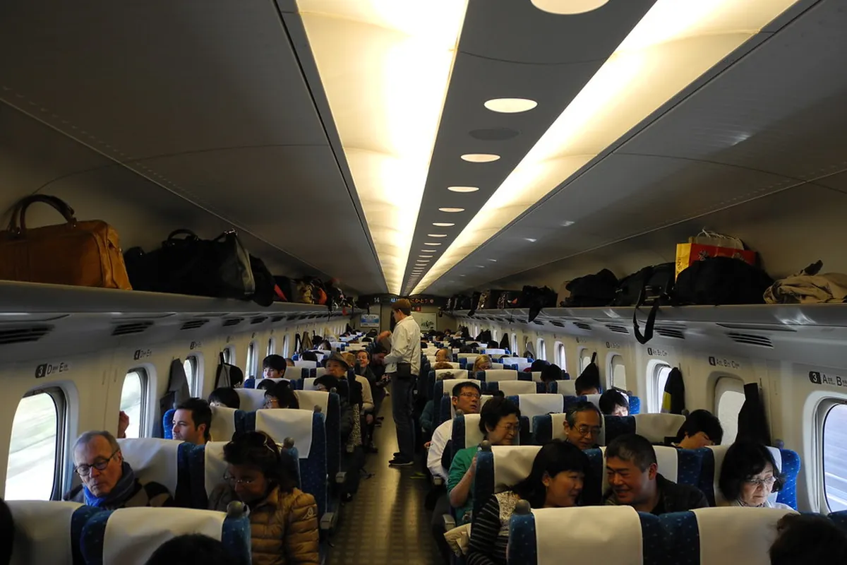新幹線の車内で、座席に座った大勢の乗客と頭上の荷物棚に置かれた荷物が写る様子の写真