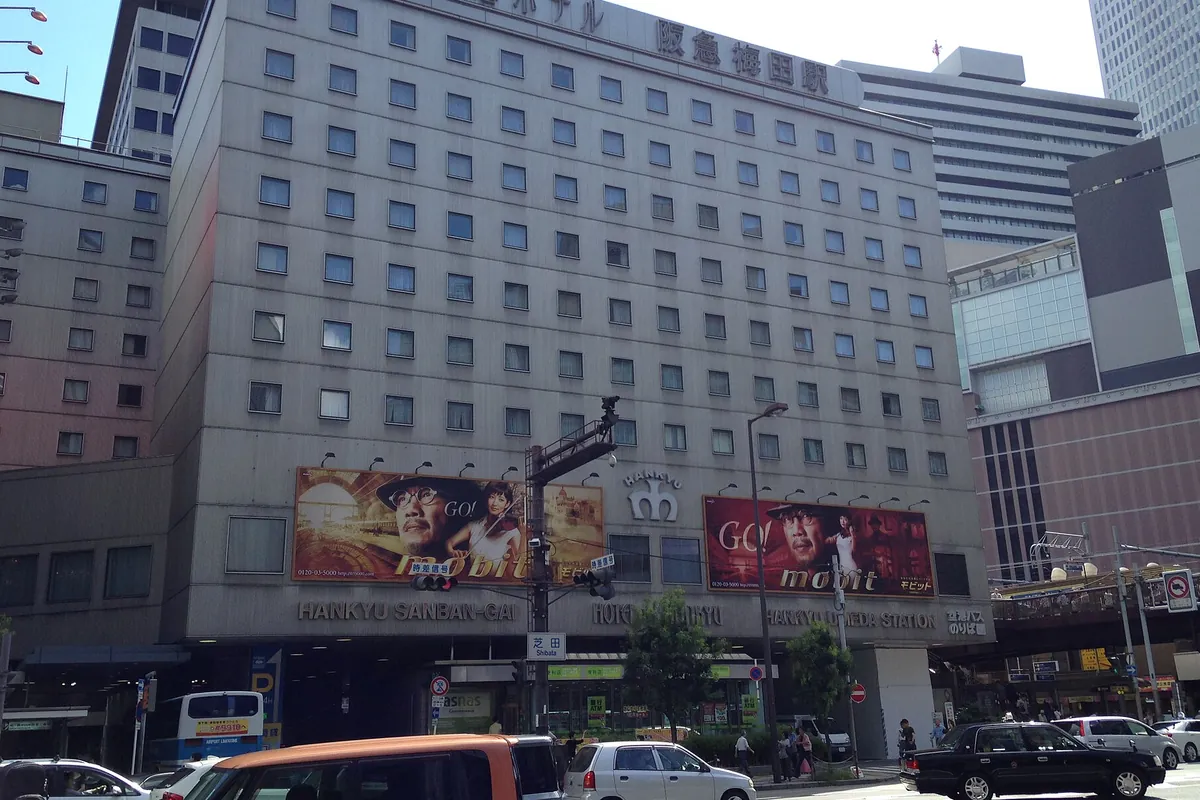 大阪梅田駅前のホテル阪急の建物に「HANKYU SANBAN-GAI」などの看板と広告が掲げられ、周辺に車やバスが行き交う様子の写真