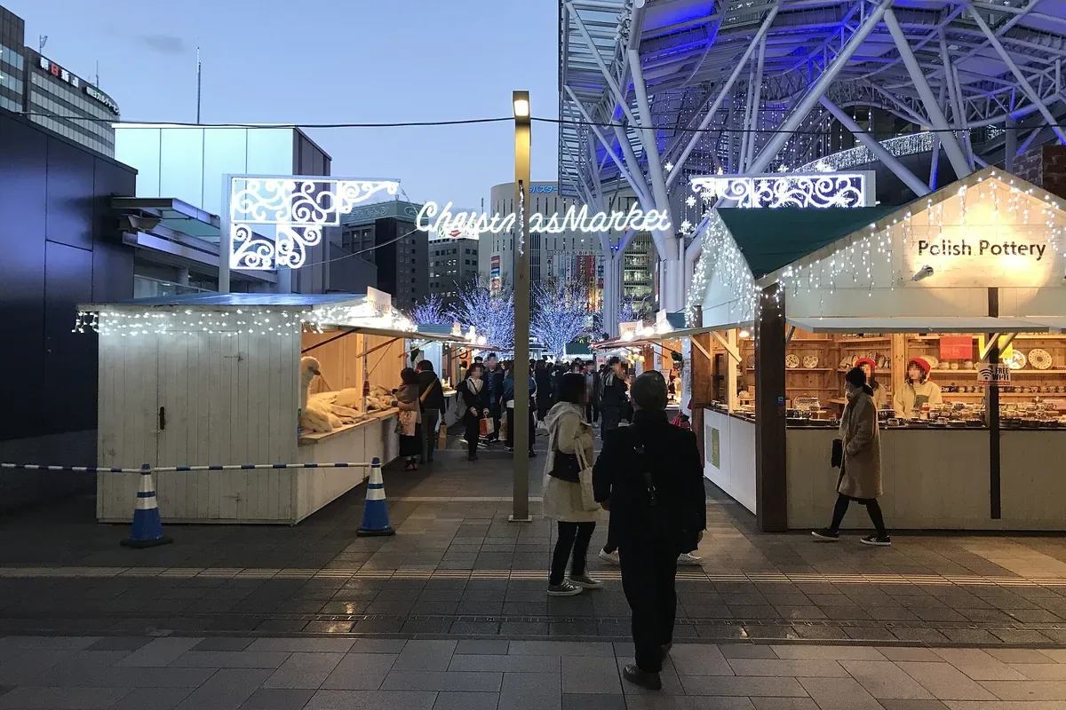 JR博多駅前の広場に白い木の小屋の店が並ぶクリスマスマーケットを、夕方に人が歩いている写真