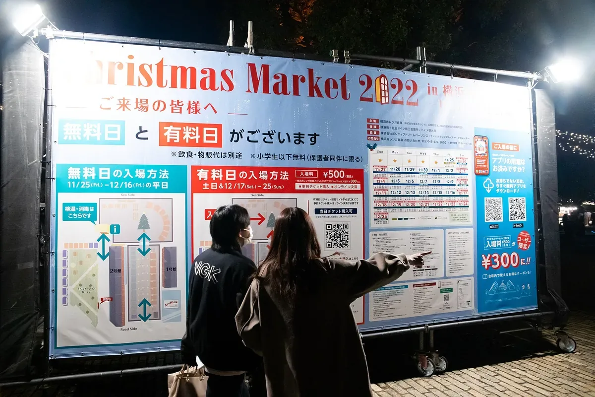 横浜赤レンガ倉庫のクリスマスマーケットの入口で、無料日と有料日の入場方法を書いた大きな案内板を2人が見ている写真