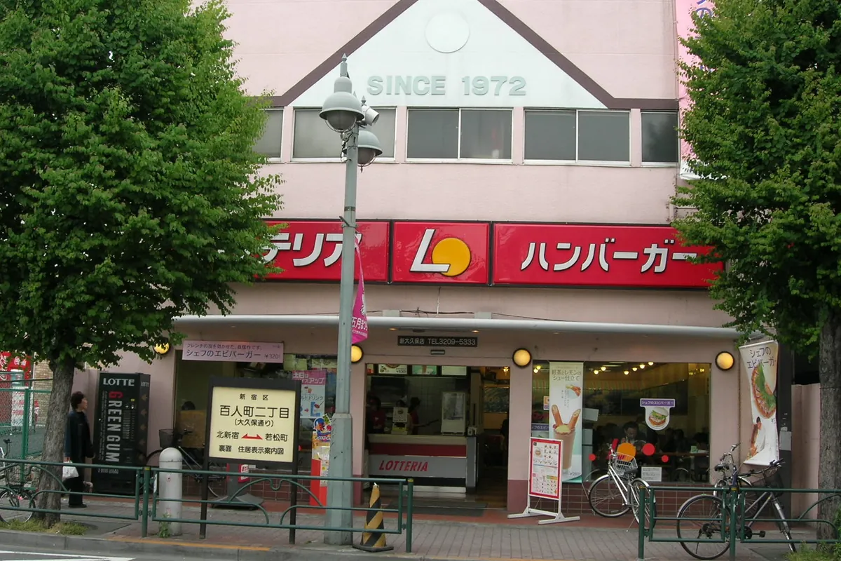 新宿区百人町の街並みで、1972年創業のハンバーガー店の店舗前に街路樹と歩道がある様子の写真