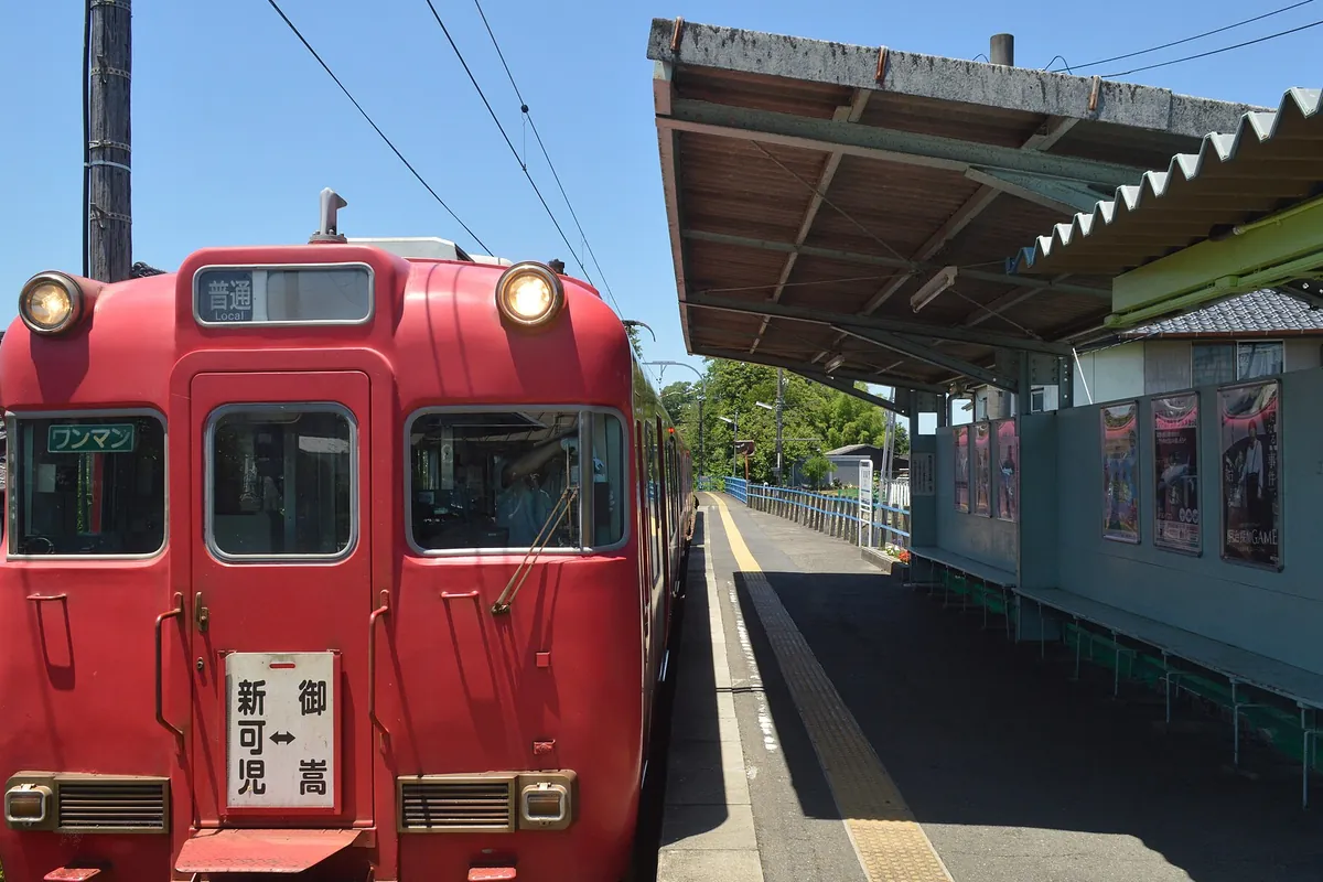 赤い車体の名鉄電車が「新可児」行きの表示でホームに停車し、屋根付きの駅のホームに広告看板が並ぶ様子の写真