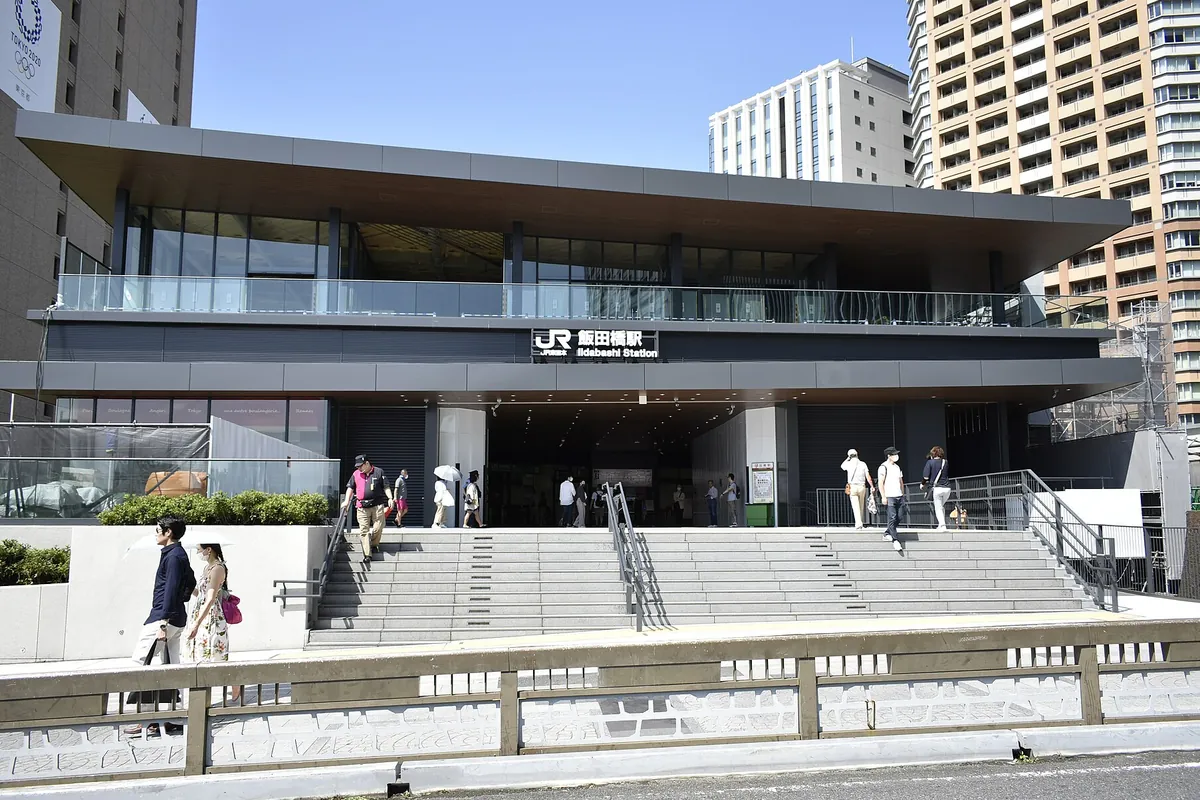 JR飯田橋駅の東口の階段と駅舎を背景に、通行人が歩道を歩いている街並みの写真