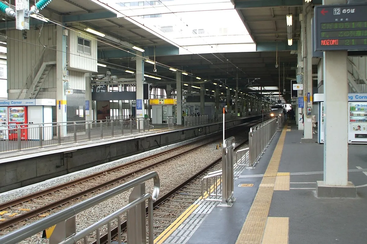 新幹線駅のホームで、架線と線路の奥から「NOZOMI」と表示された新幹線が近づいてくる様子の写真