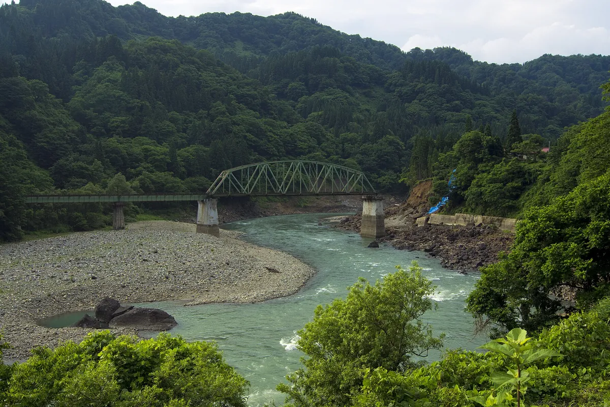 緑の山に囲まれた渓谷を流れる川に架かるトラス構造の鉄橋を見下ろした写真