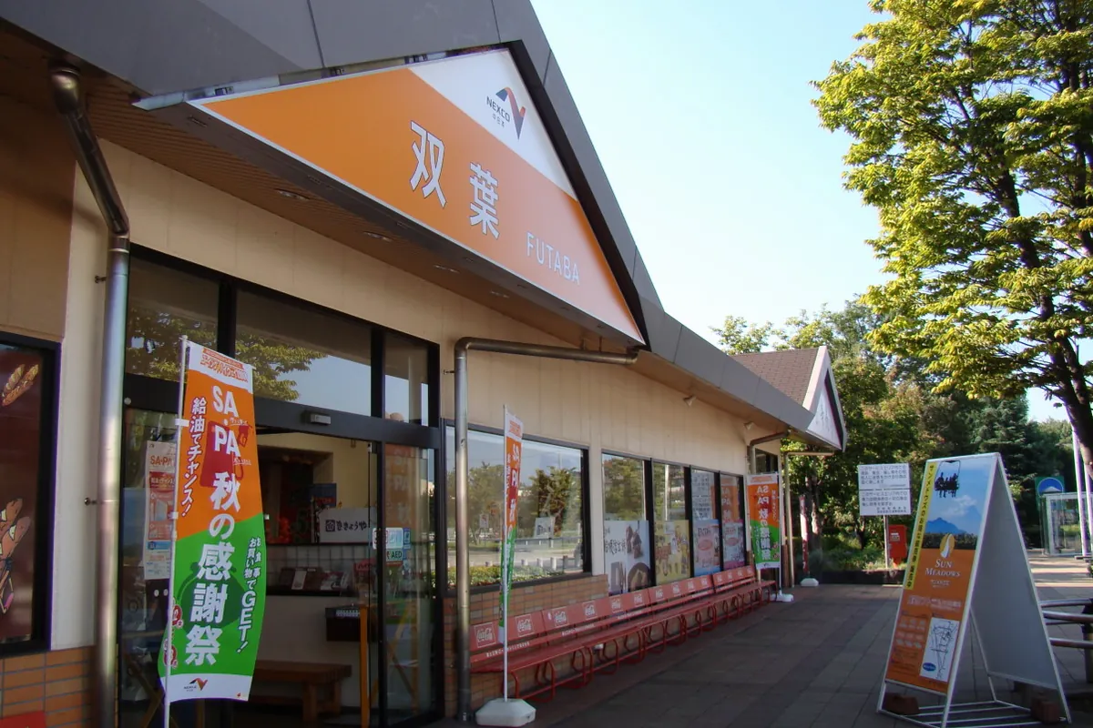 サービスエリアの建物に「双葉」と書かれた看板が掲げられ、入口前にベンチやのぼりが並ぶ様子の写真