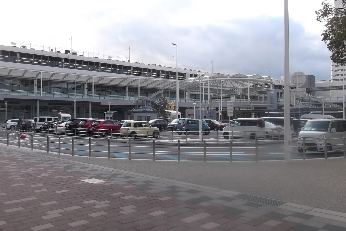 駅前ロータリーに面した駅ビルの建物と、手前に並んで駐車する多数の乗用車の様子の写真
