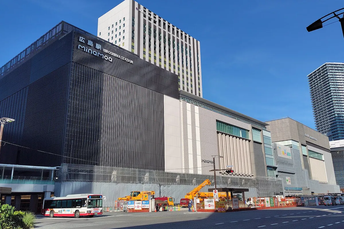 広島駅ビル「HIROSHIMA STATION MinamOa」の看板が掲げられた外観と、手前にバスや作業車が並ぶ様子の写真