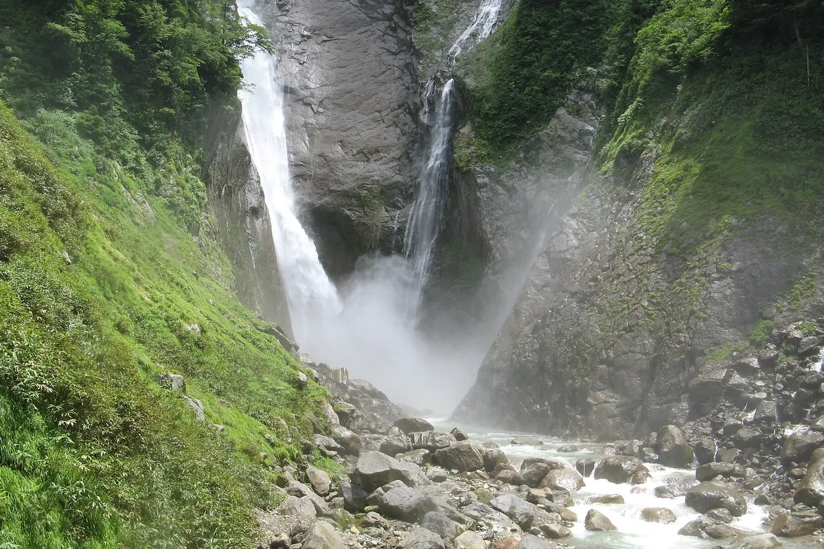 険しい岩壁を二筋に分かれて流れ落ちる称名滝と、その下の川に転がる岩の様子の写真