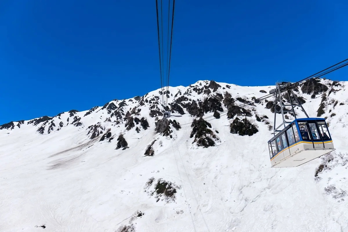 雪に覆われた立山連峰の斜面を背景に、立山ロープウェイのゴンドラがケーブルで運行している様子の写真