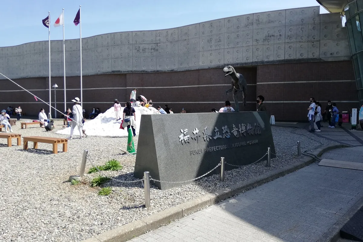 福井県立恐竜博物館の入口前で、恐竜の像と館名が刻まれた石碑が立ち来場者が歩く様子の写真