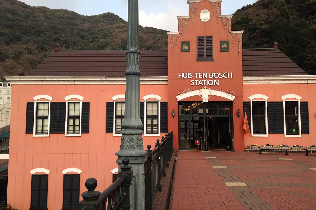 「HUIS TEN BOSCH STATION」と書かれたオレンジ色の駅舎の正面に、装飾的な街灯と煉瓦敷きの広場が広がる様子の写真