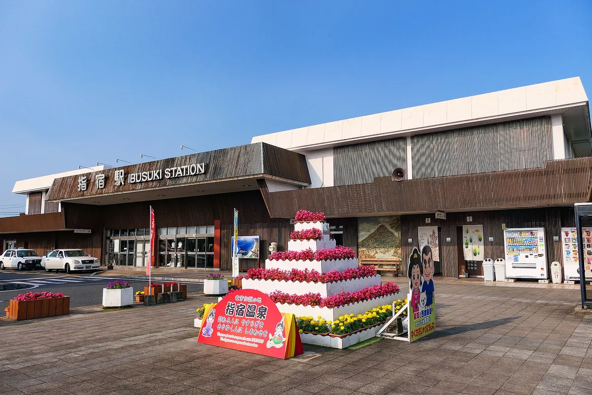 花壇の飾り付けがある広場の奥に、「指宿駅 IBUSUKI STATION」と表示された駅舎が写る写真