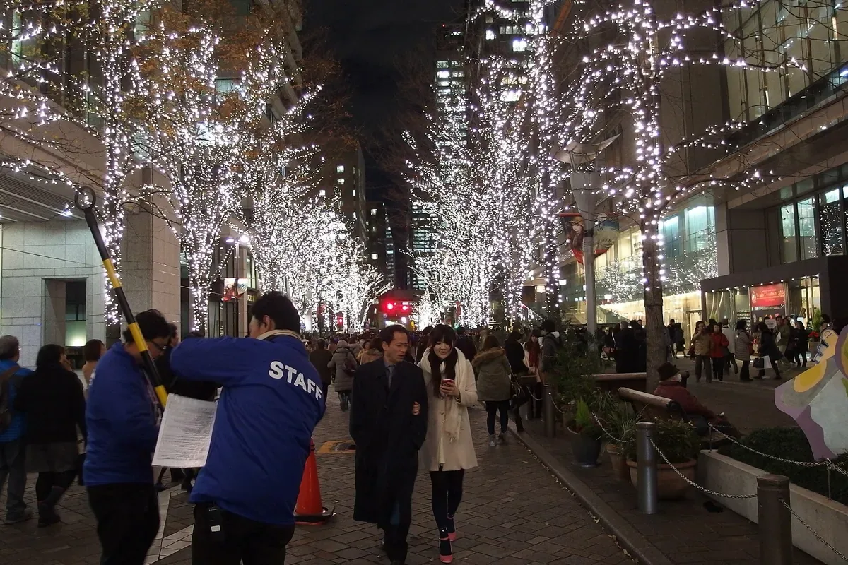 クリスマスイブの丸の内仲通り。白く光る並木の下の歩行者の道に人が集まり、係の人が案内の看板を持って立っている夕方の写真