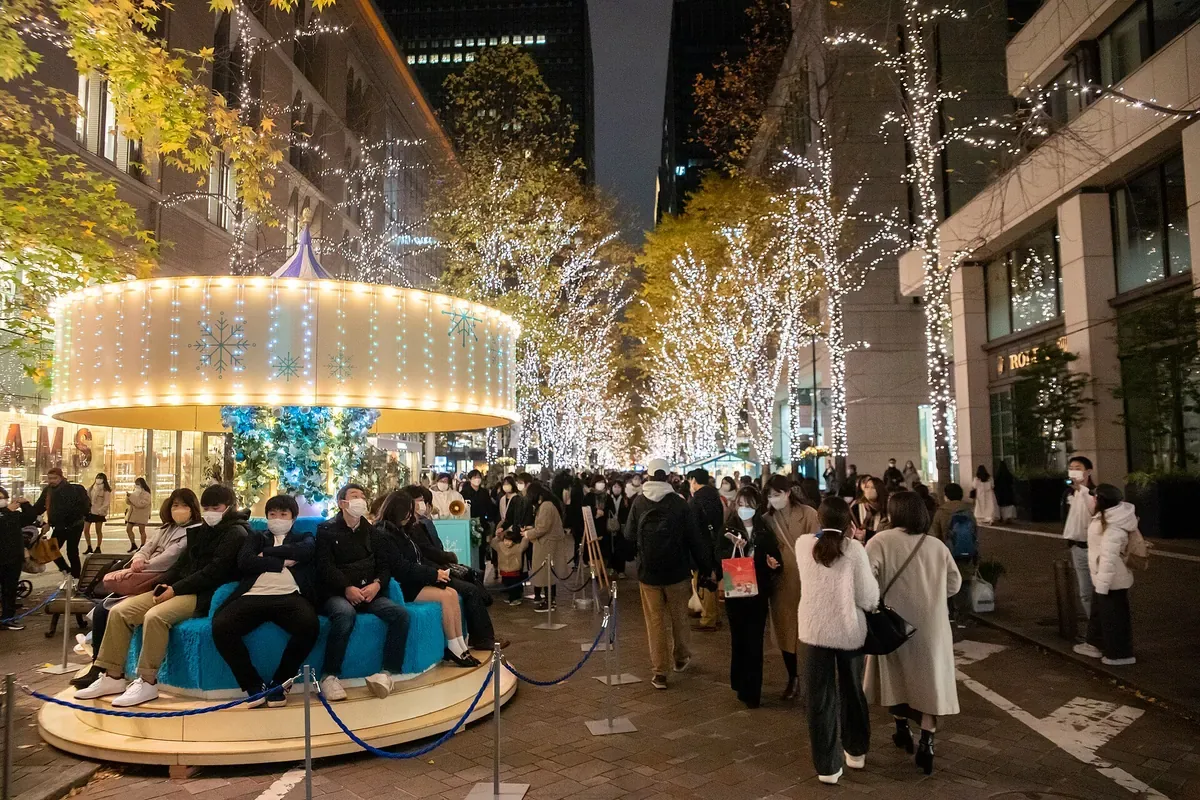 丸の内仲通りのイルミネーション。白い光をまとった並木の下を大勢の人が歩き、手前の光のメリーゴーラウンドに人が腰かけている夜の写真