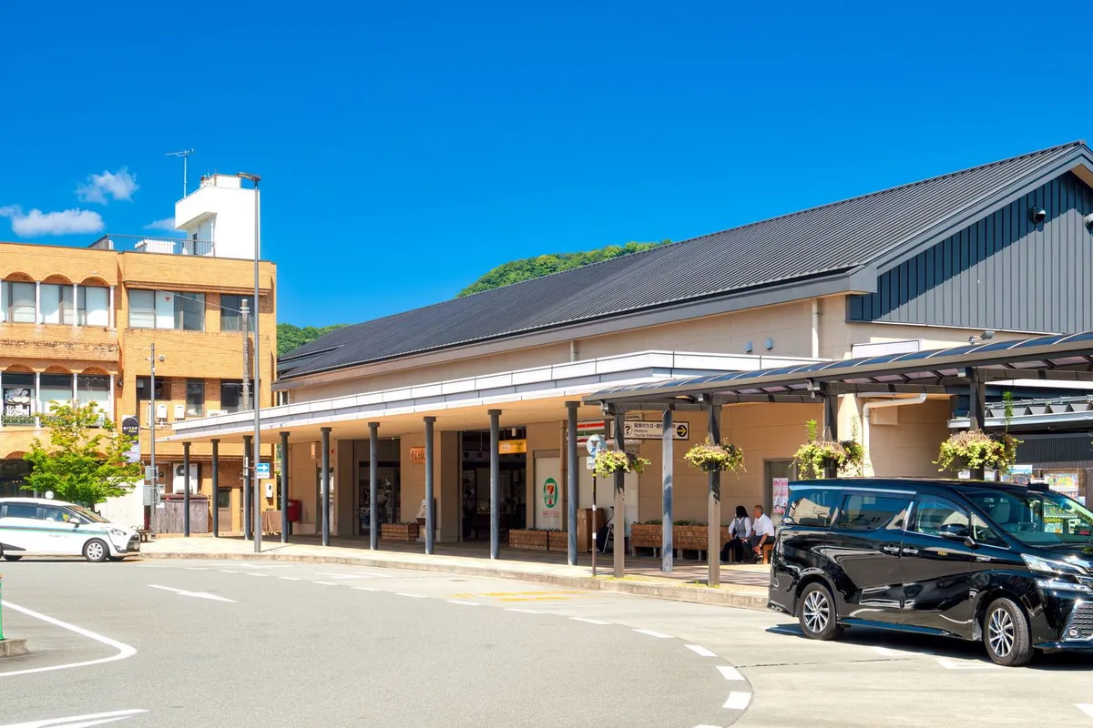 駅前の広場に黒い車やタクシーが停まり、瓦屋根の駅舎とセブンイレブンの看板が並ぶ写真