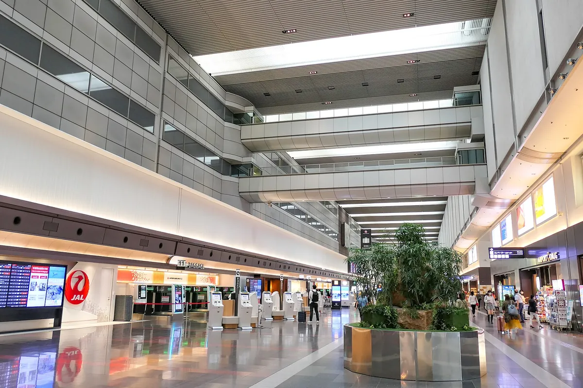 空港の出発ロビーでJALのロゴが掲げられたチェックインカウンター前を利用客が行き交う様子の写真
