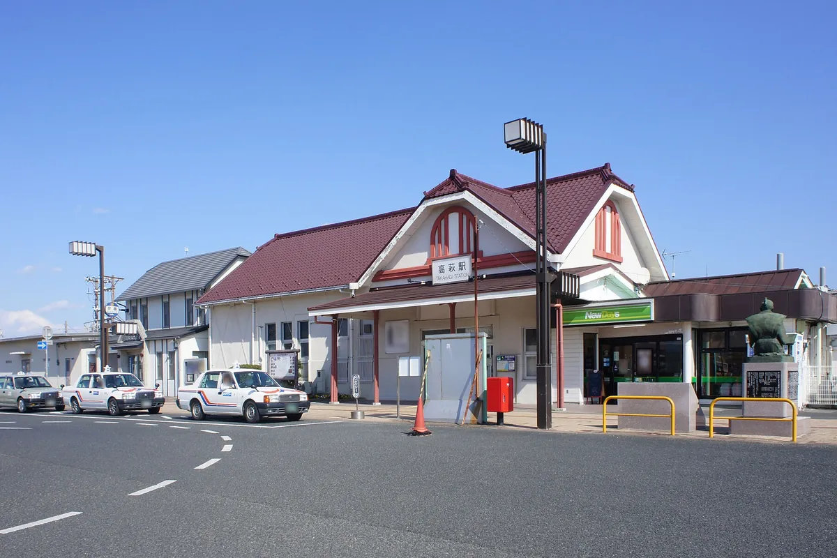 タクシーが並ぶロータリーの奥に、赤い三角屋根と「高萩駅」の駅名表示がある木造駅舎が建つ様子の写真