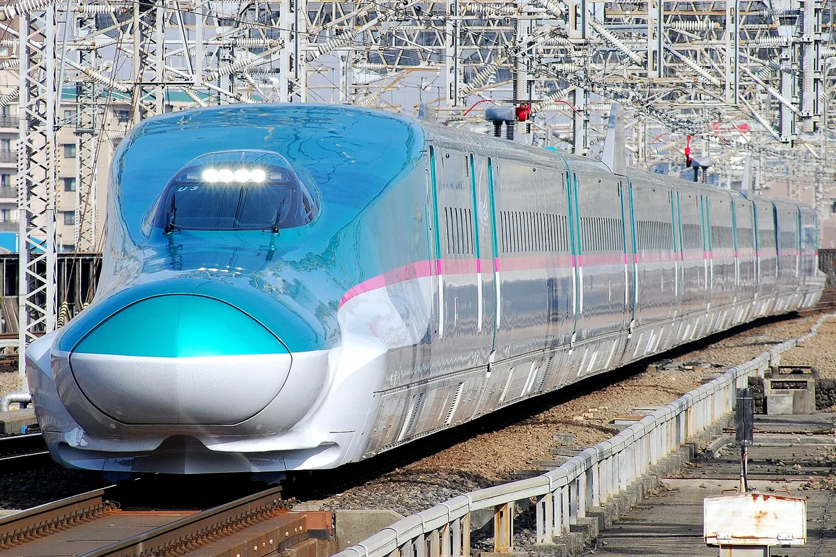 緑色と白を基調としたE5系新幹線車両が架線の鉄塔や高架設備が並ぶ線路を走行している様子の写真