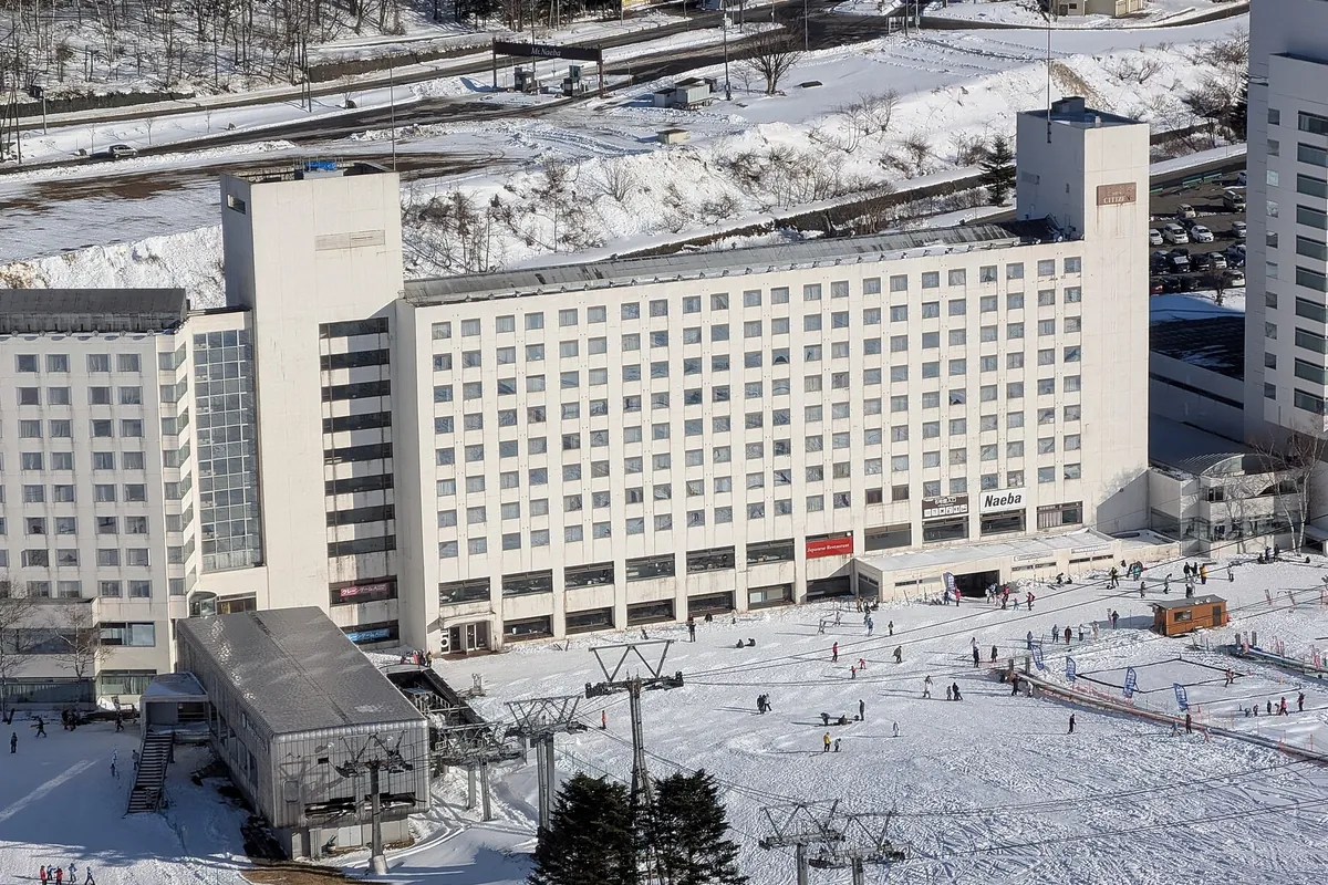 雪に覆われたゲレンデと「Naeba」の表示がある大型ホテル建物、スキー客が行き交う様子の写真