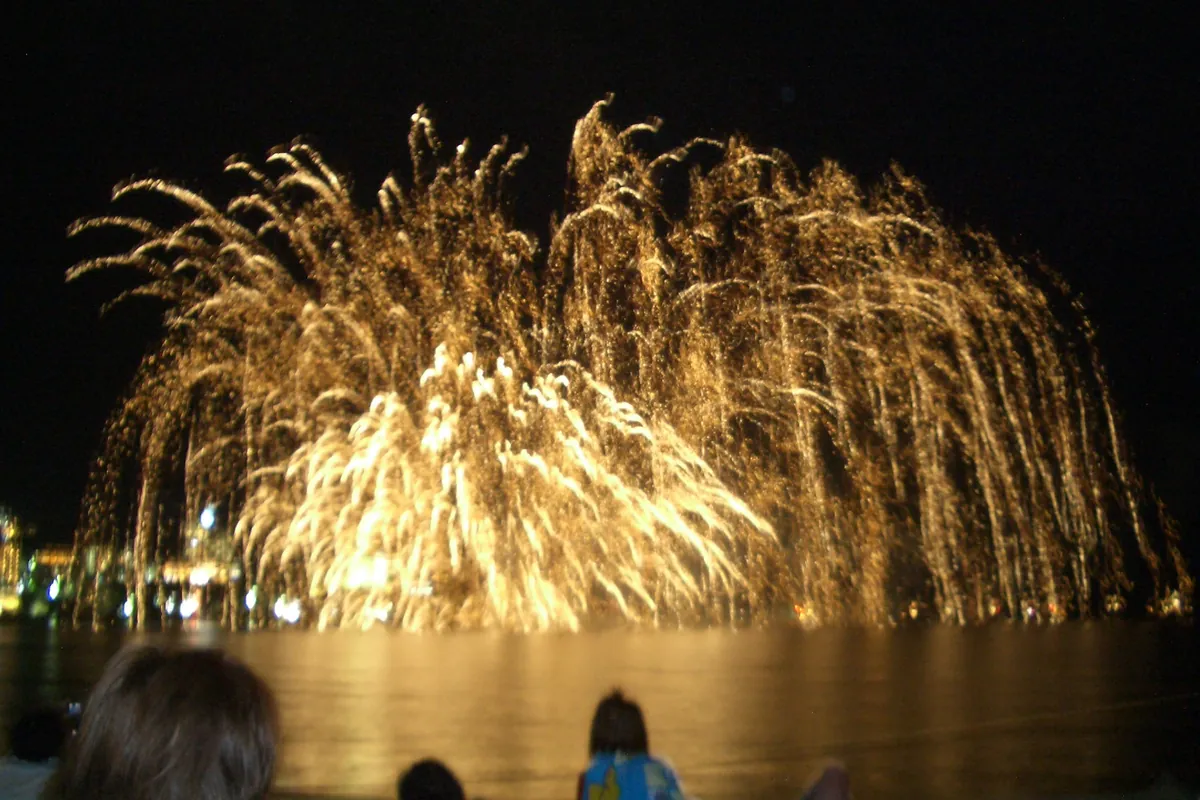 水辺の花火大会で、大きく開いた金色の花火と見物する人の後ろ姿が写る夜景の写真