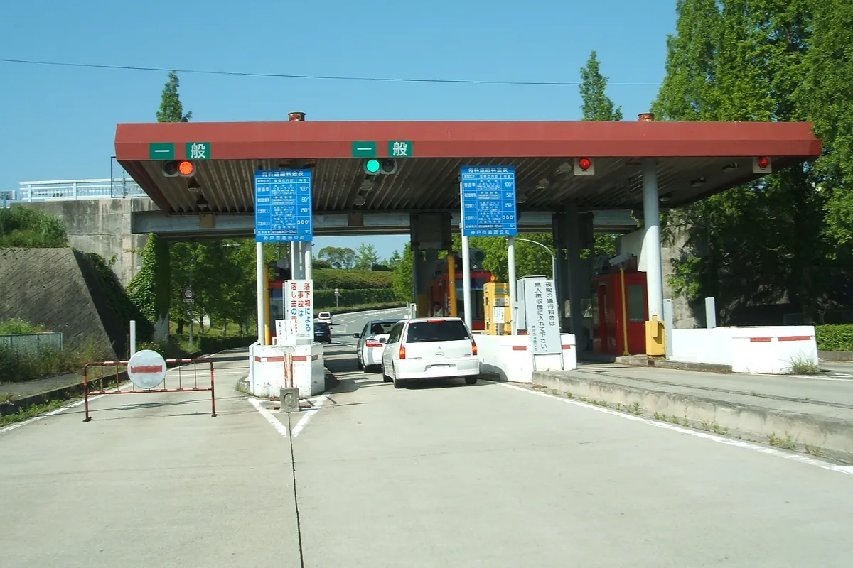 有料道路の料金所で、通行料金の案内板や遮断機、通過する車が写る様子の写真